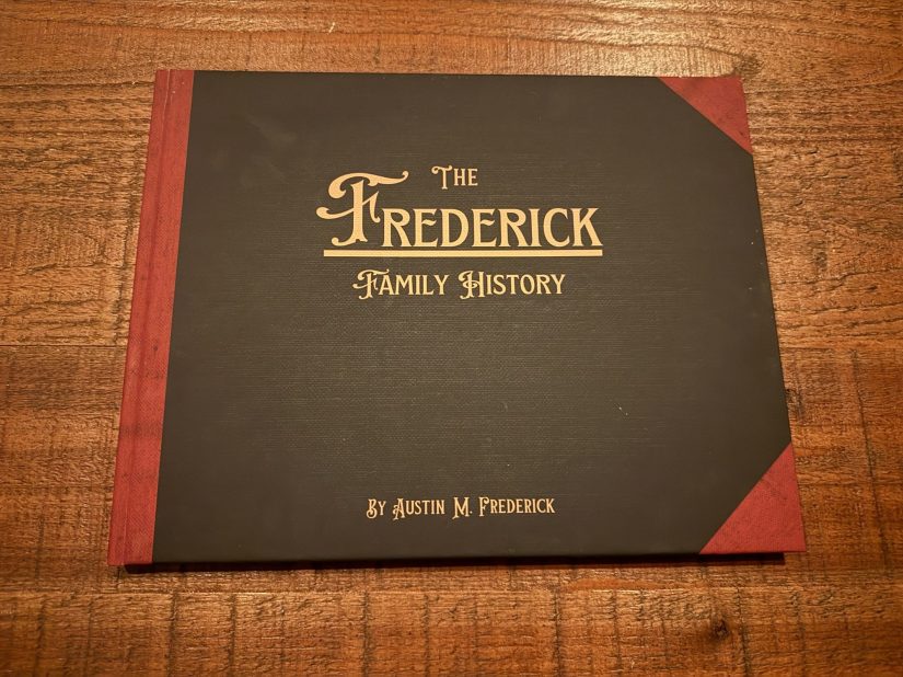 A family history book titled 'The Frederick Family History' by Austin M. Frederick, featuring a decorative cover with gold lettering and red accents on a wooden surface.